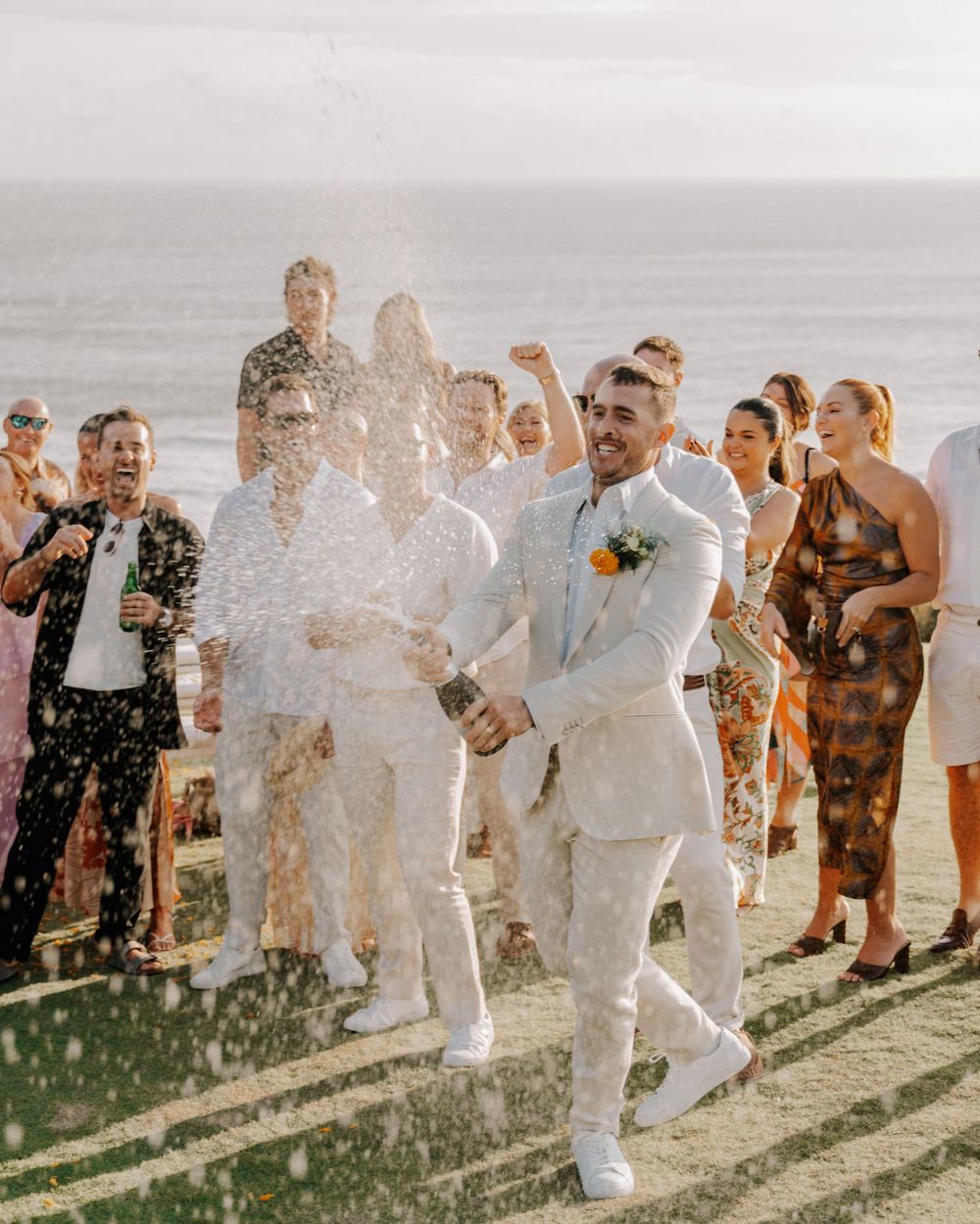 Wedding party celebrating with champagne spraying on the beach at sunset during a joyful outdoor celebration.