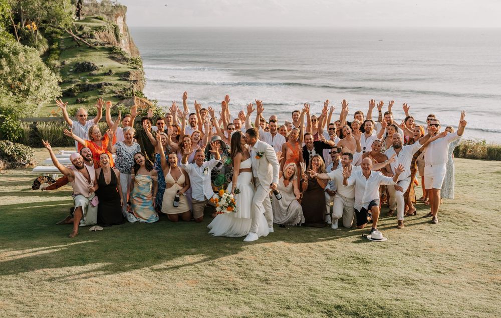 Large wedding party celebrates joyfully together on grassy lawn at sunset while raising their arms in unison.