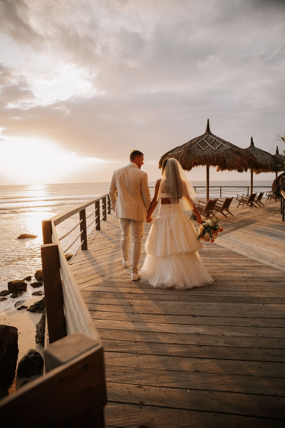 Couple in white attire walks along a wooden pier at sunset overlooking a tropical ocean view with palm umbrellas.
