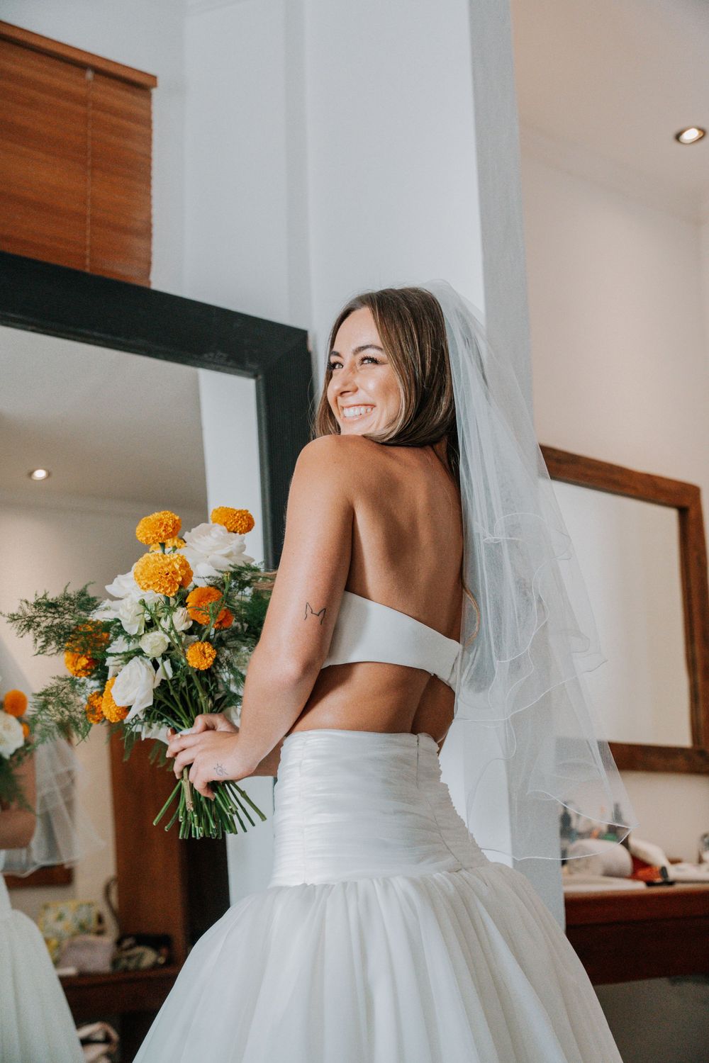 A bride in a white wedding gown holds a colorful bouquet with orange and white flowers against a bright interior.