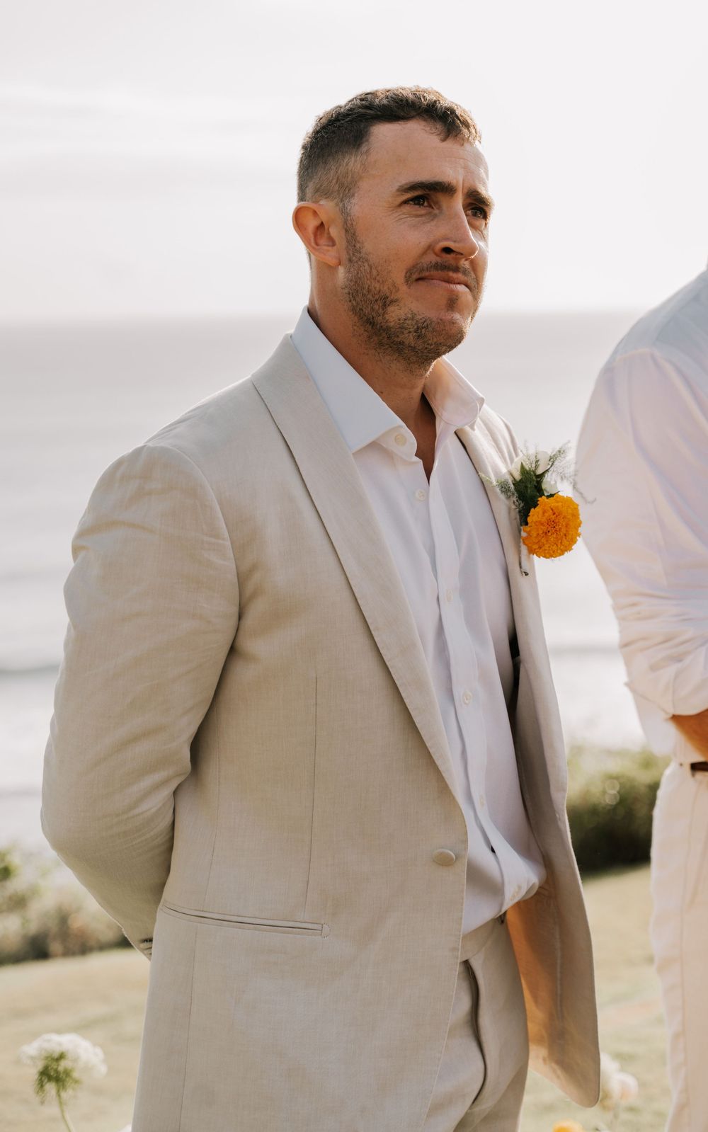 A groomsman in a beige suit and white shirt poses with an orange boutonniere during an outdoor wedding.