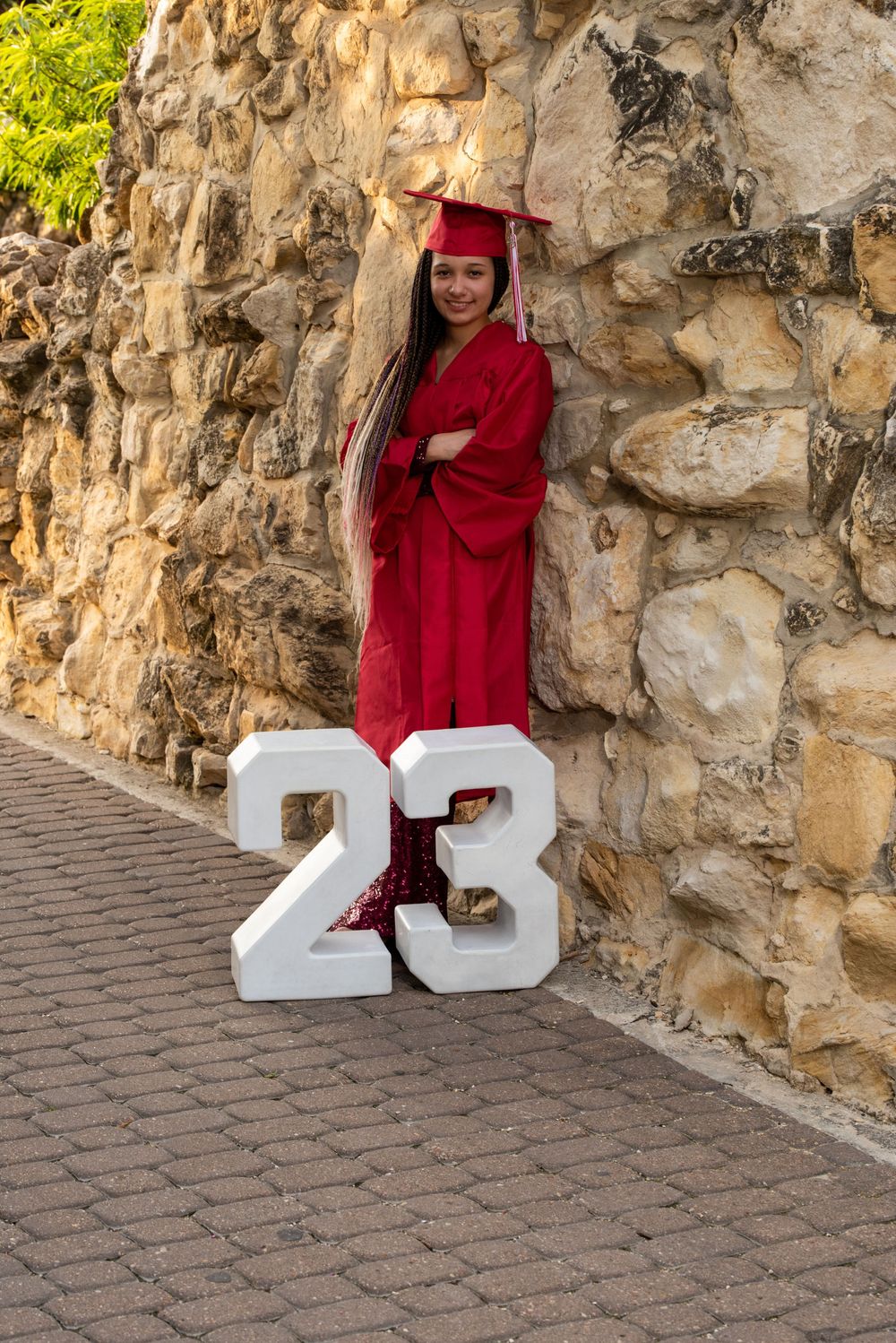 A graduate in red cap and gown poses with white number 23 decorations against a stone wall on graduation day.