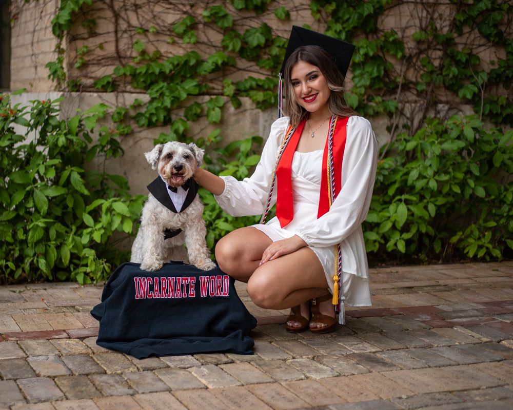 Smiling graduate in red and white attire poses on brick patio with small dog dressed in graduation cap and gown.