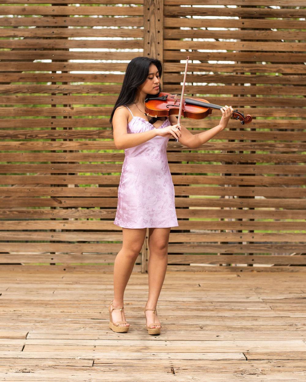 Musician in a pink dress plays violin while standing on wooden deck against slatted wood wall background.