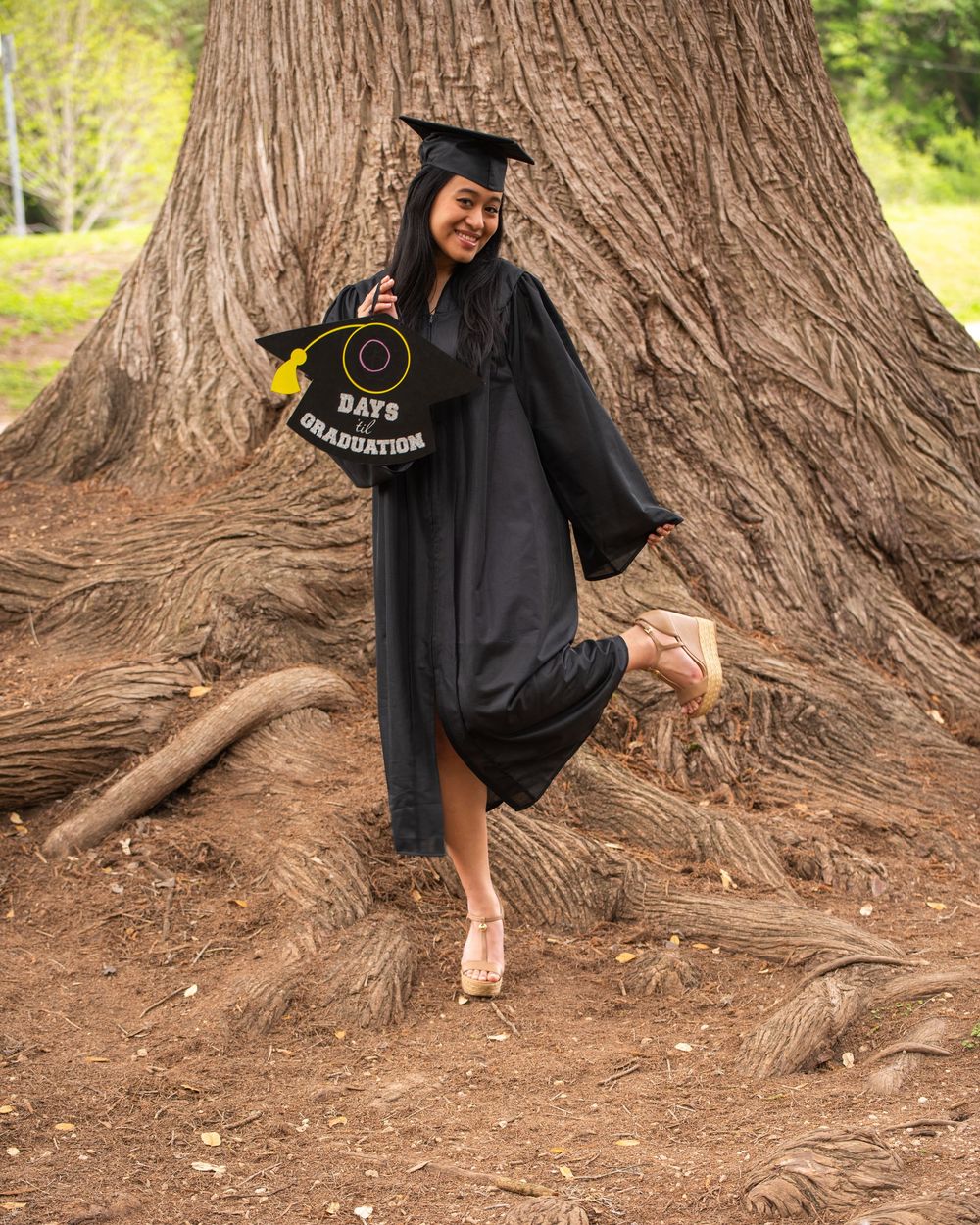 Student wearing traditional black graduation gown poses playfully at base of large tree trunk in outdoor setting.