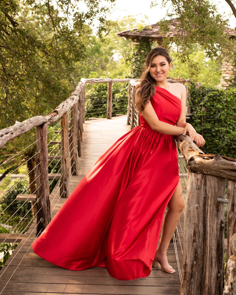 Woman in flowing red dress poses gracefully on rustic wooden deck surrounded by lush greenery.