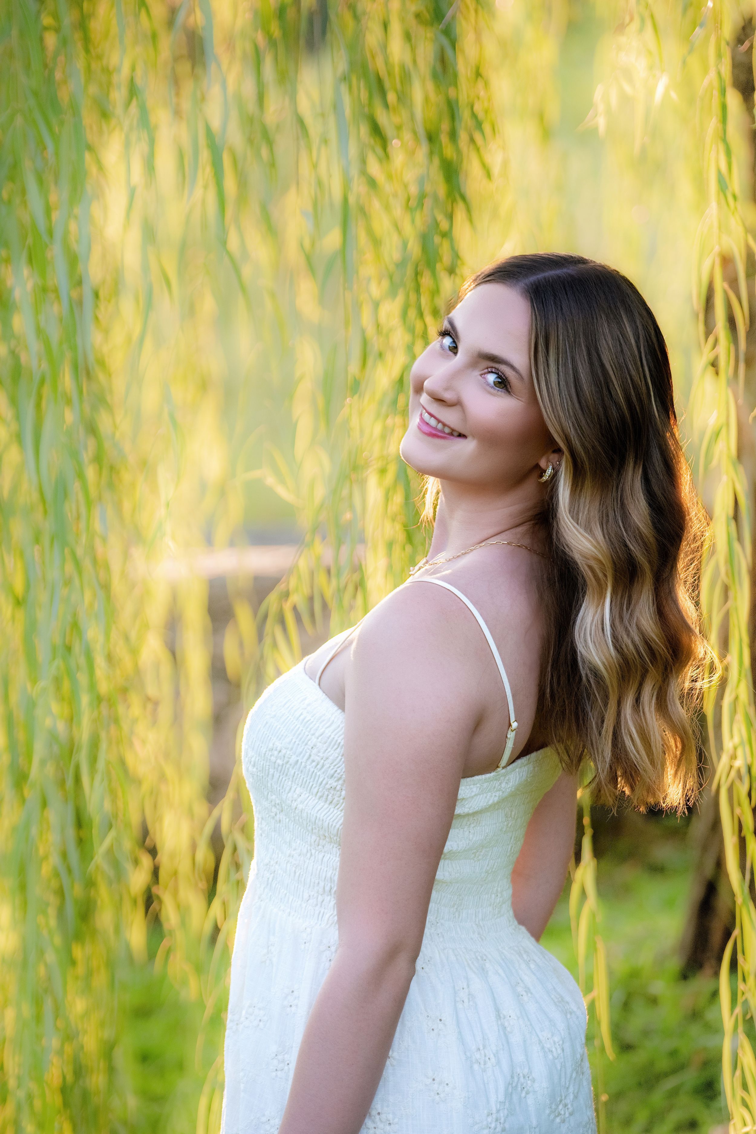 A young woman in a white summer dress poses gracefully in front of cascading willow branches in warm golden light.