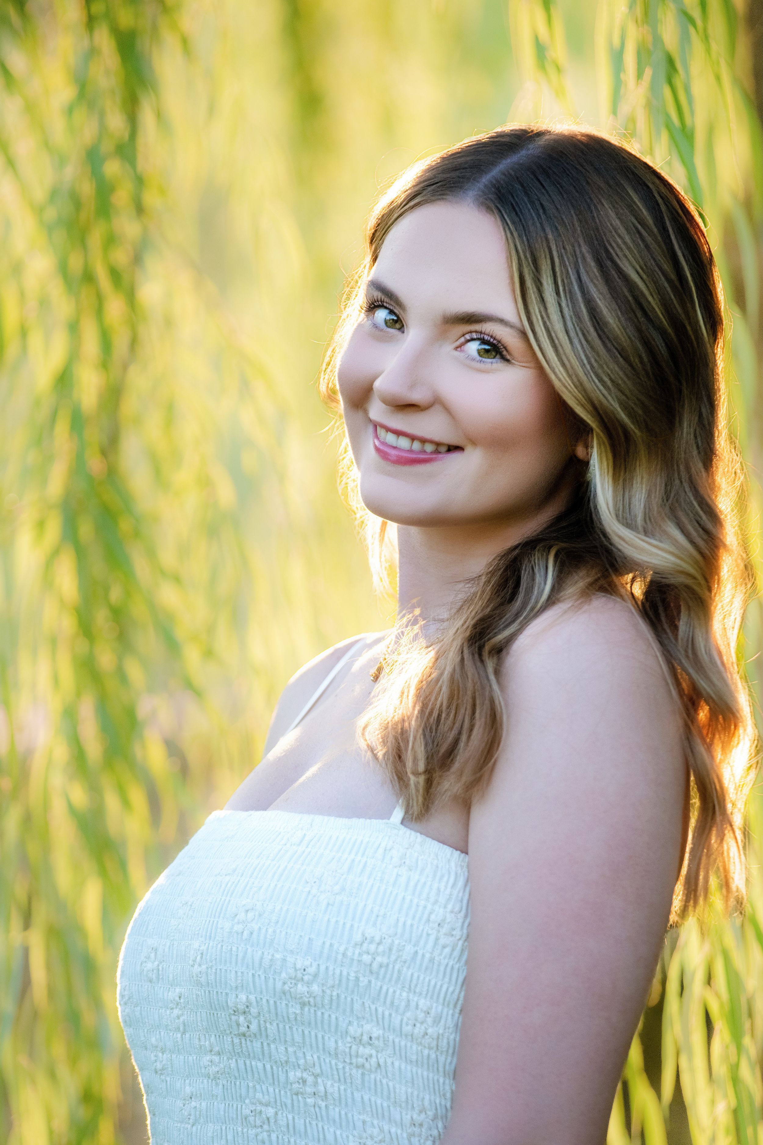 A person in a white sleeveless dress poses outdoors with a backdrop of sunlit willow branches on a sunny day.