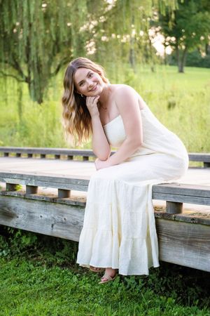 A person in a flowing white dress poses gracefully on rustic wooden steps surrounded by lush green summer foliage.