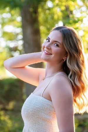 A smiling woman in a white top poses in natural sunlight with lush greenery and bokeh lighting in the background.