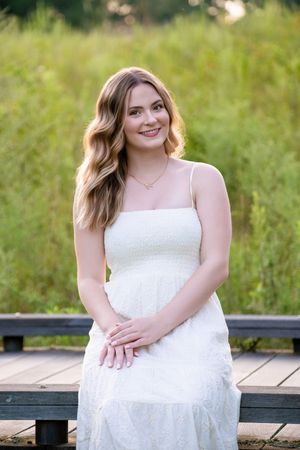 A woman in a flowing white sundress sits on a wooden deck surrounded by greenery on a sunny day.