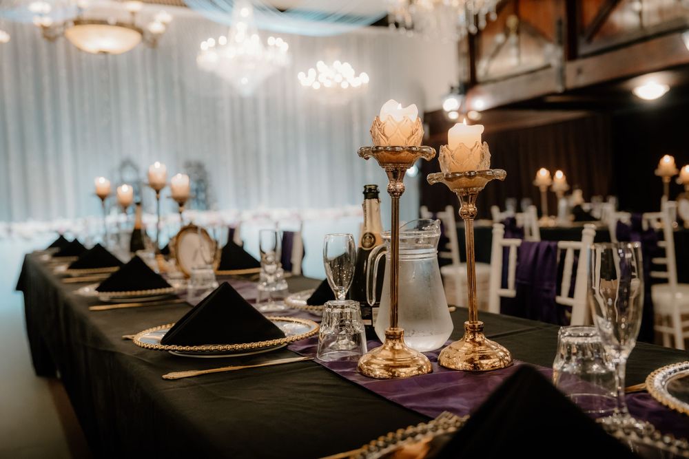 Elegant wedding reception table with gold candelabras, black napkins, purple runner, and crystal glassware in a decorated venue.