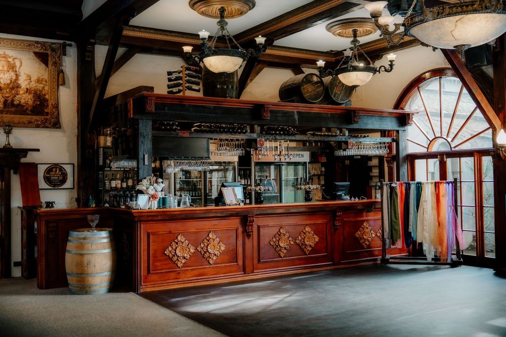 Rustic winery tasting room with ornate wooden bar, barrel decor, chandelier, and colorful scarves near arched window.