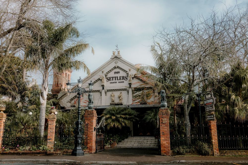 Settlers Country Manor historic building entrance with ornate iron gates, brick pillars, and lush palm trees.