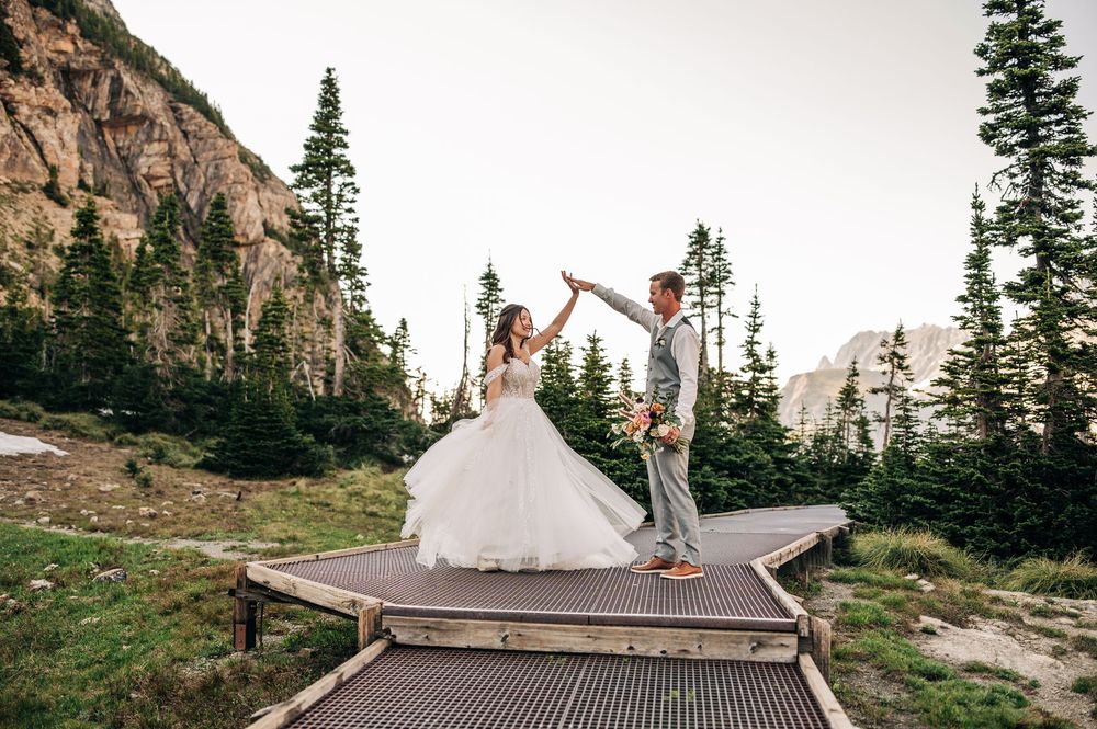 A couple in wedding attire poses joyfully against the backdrop of majestic mountain peaks at sunset.