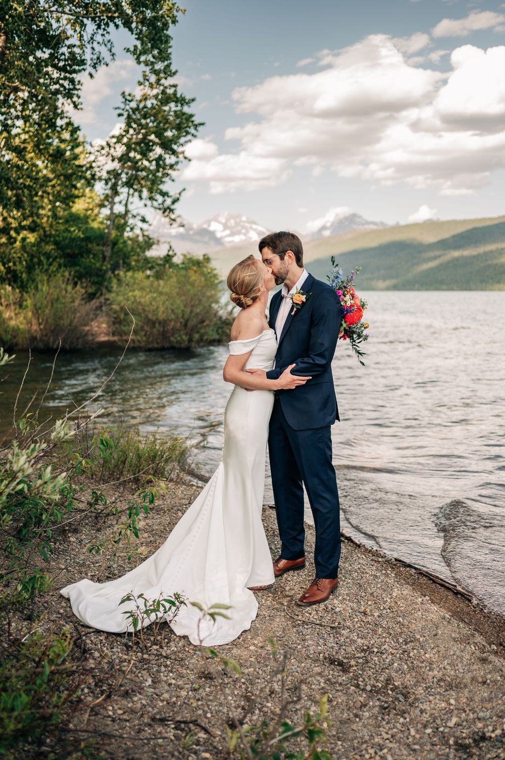 A couple shares an intimate moment on a rocky lakeshore with mountains and cloudy skies in the background.