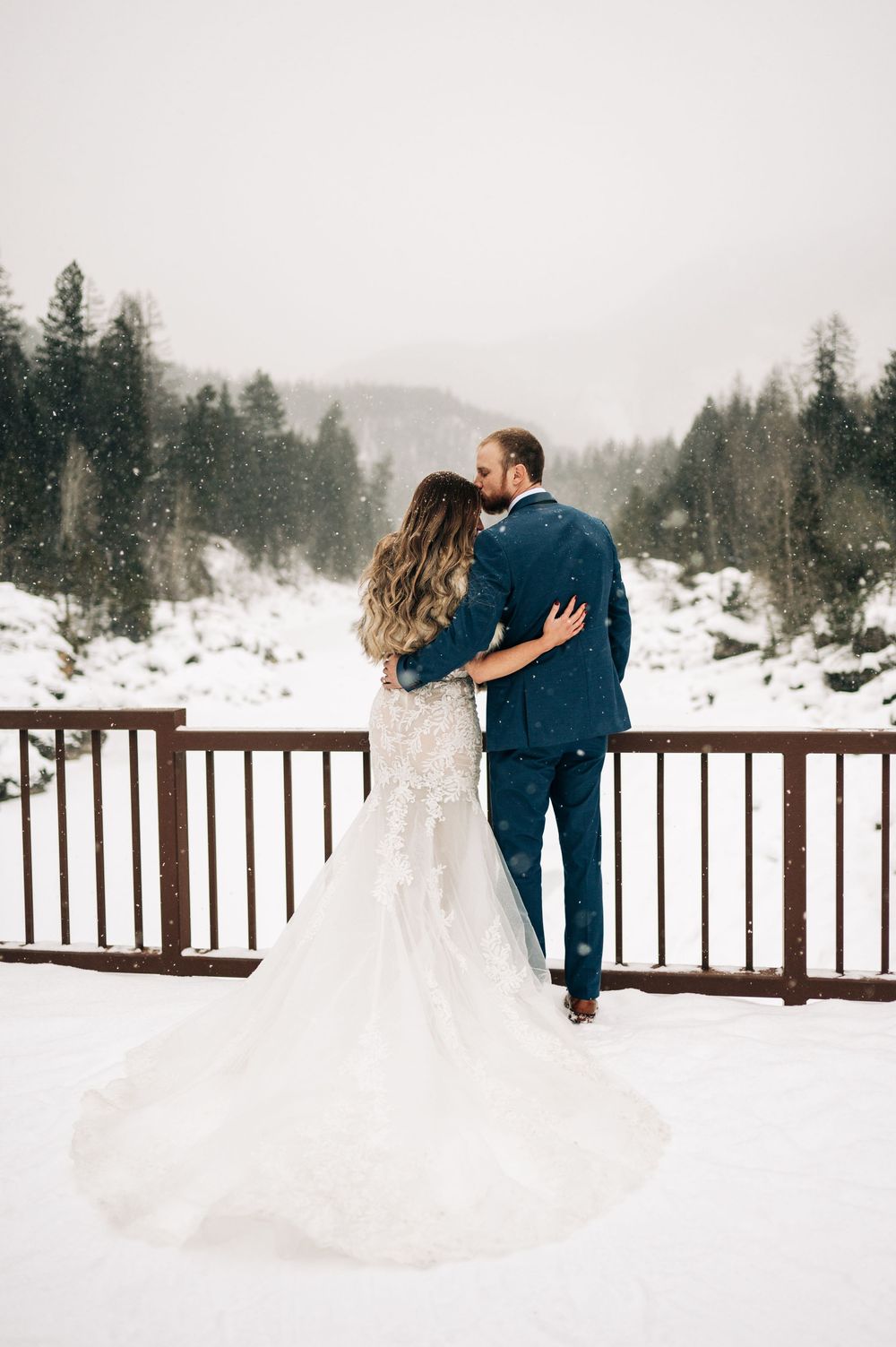 A couple in wedding attire embraces on a snowy deck overlooking a serene winter landscape at their mountain ceremony.