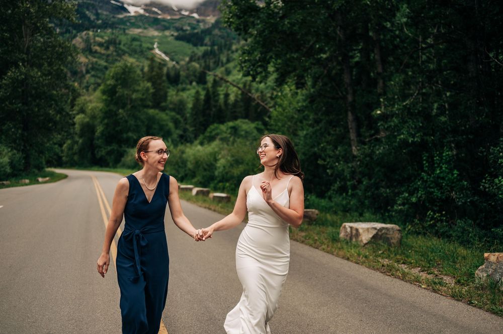 A couple in wedding attire walk hand in hand along a winding country road while sharing joyful laughter together.