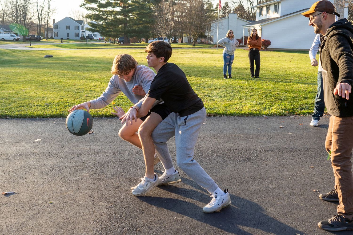 Group of youths play basketball on a driveway with others gathered to watch.