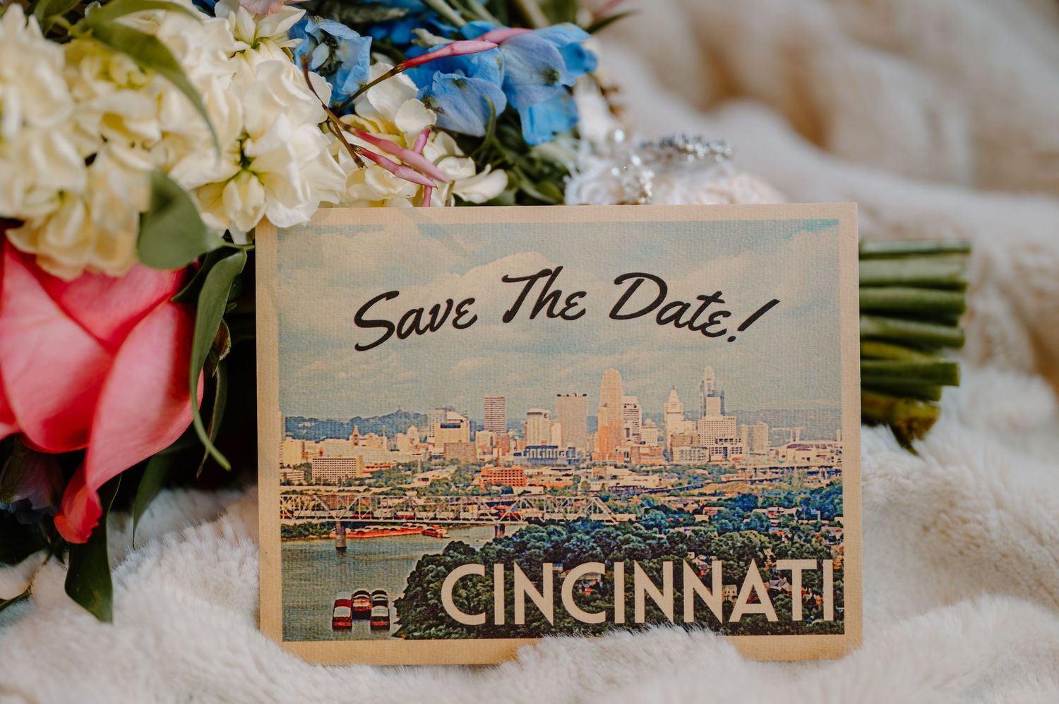 A postcard showing a panoramic skyline view of downtown Cincinnati with text 'Save the Date' on a table with floral decor.