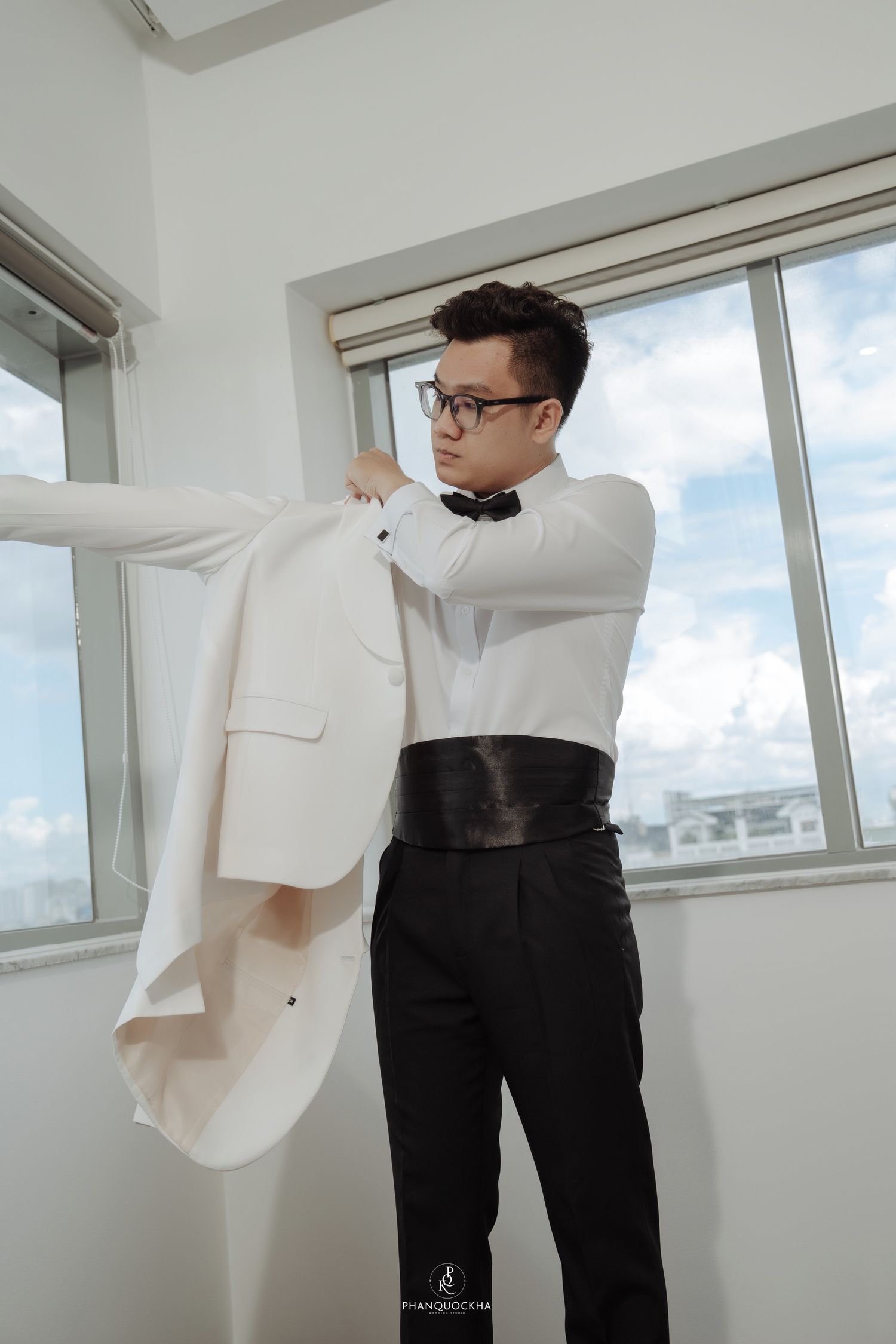 Asian groom putting on white tuxedo jacket with black bow tie and cummerbund by windows.