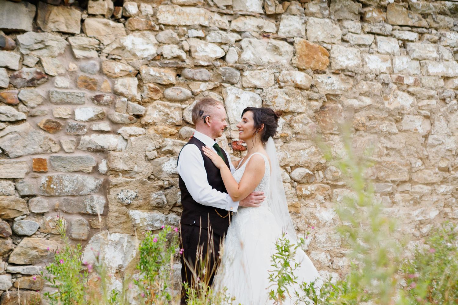 Tender embrace against weathered stone wall with wildflowers in foreground.