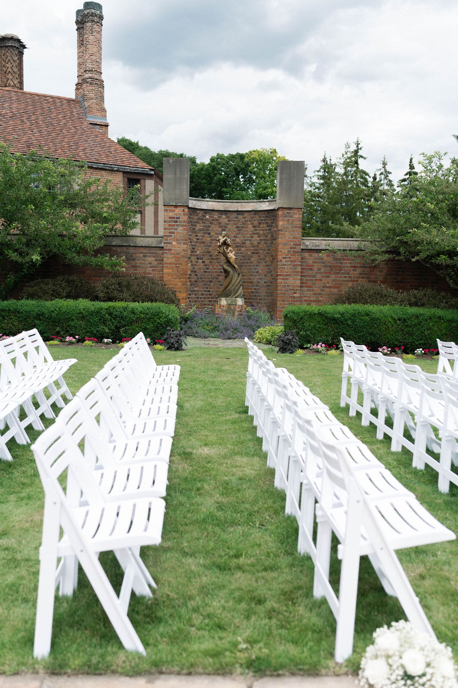 Rows of white wedding chairs set up on lawn in front of brick wall with decorative fountain.