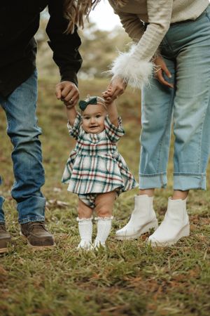 Baby in plaid dress taking first steps between parents wearing jeans.