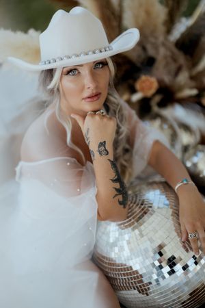 Woman in western attire poses with disco ball and shows tattoo art in dreamy setting.