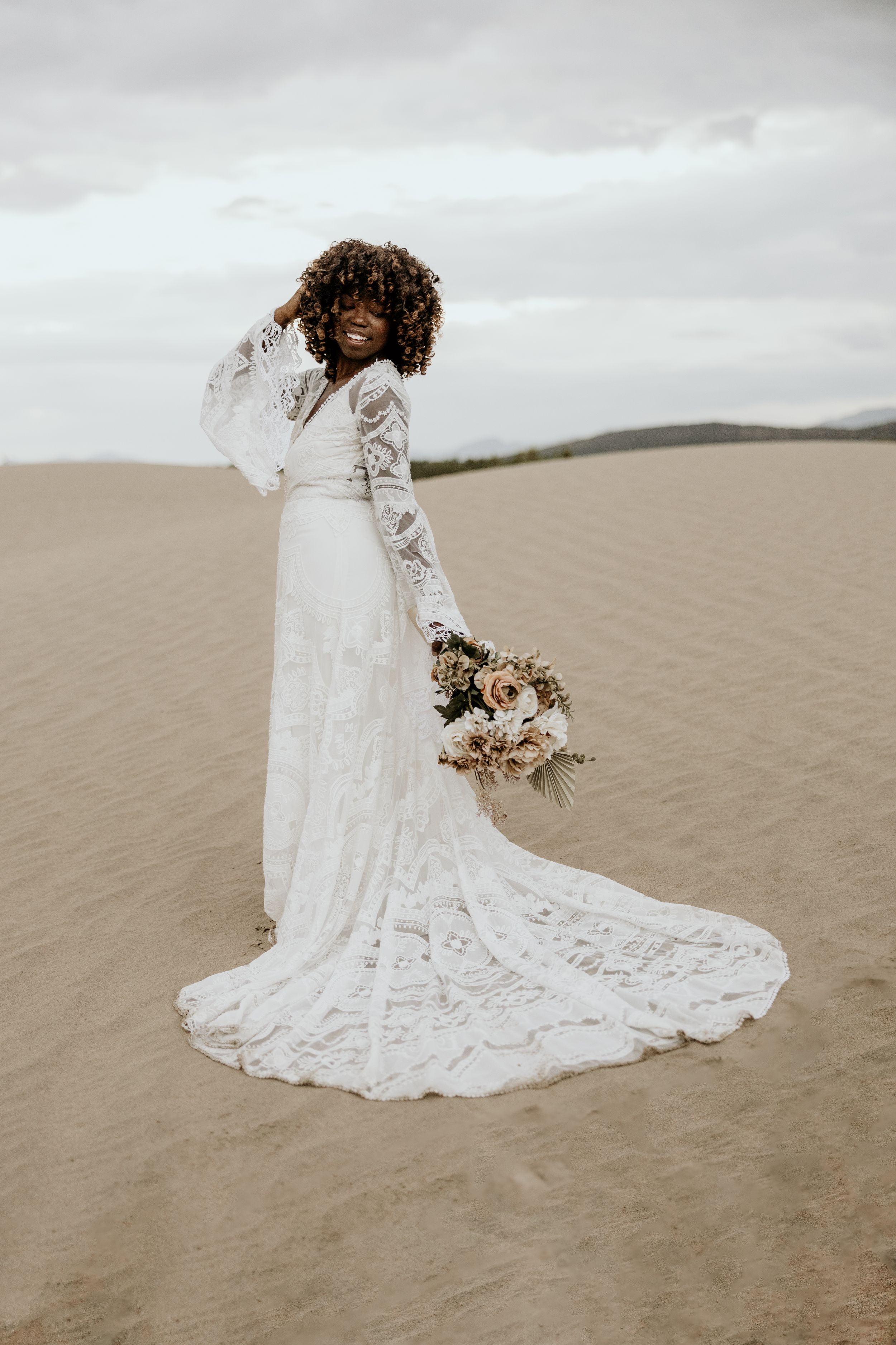 Bride in long-sleeve lace wedding gown holding bouquet poses on sandy dunes under cloudy sky.