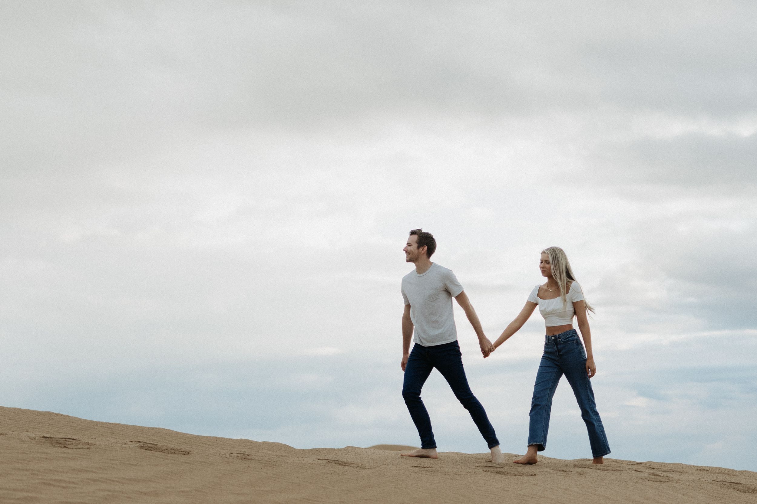 Couple holding hands while walking on sand dunes against cloudy sky.