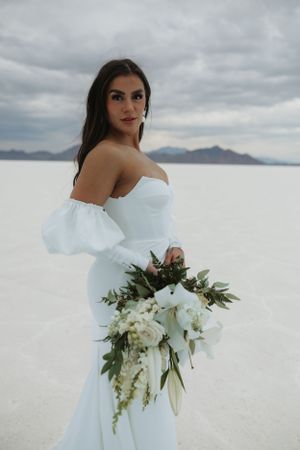 A bride in an elegant white gown holds a lush bouquet against stark white salt flats.