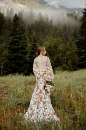 A bride in a detailed lace gown stands against a misty mountain forest backdrop.