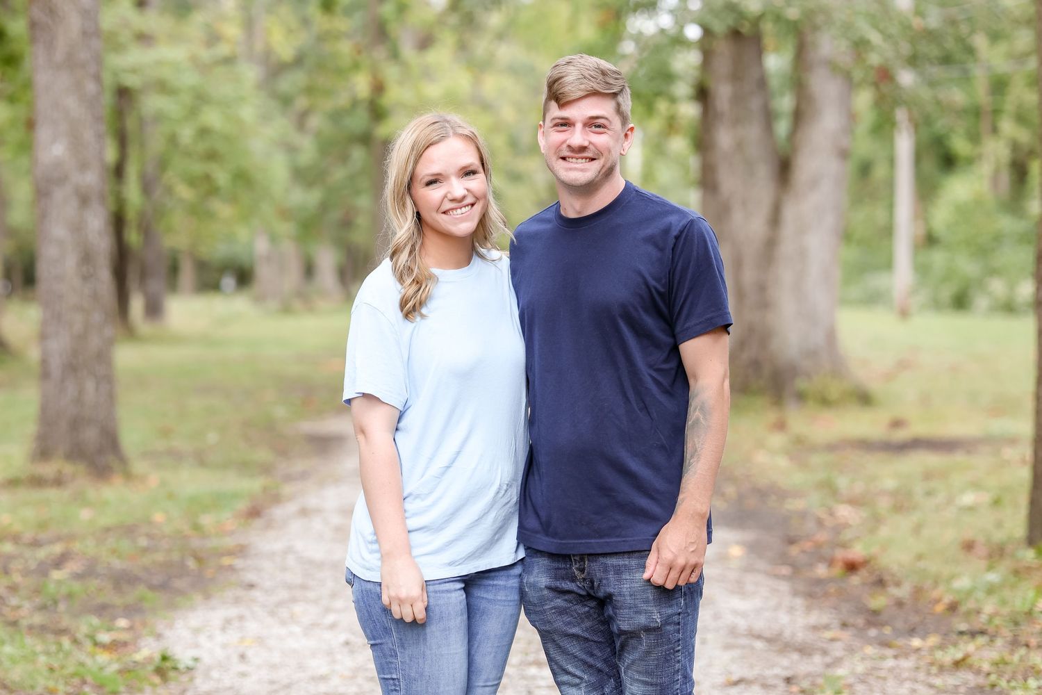 Casually dressed siblings stand together on a wooded path, smiling at the camera on a sunny day.