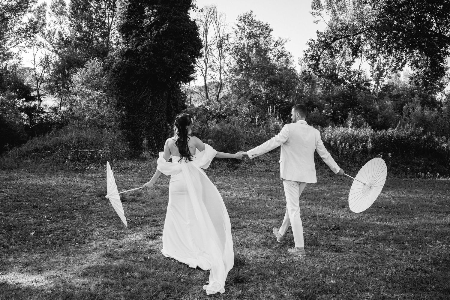 Bride and groom hold hands while spinning with parasols in a lush green outdoor setting.