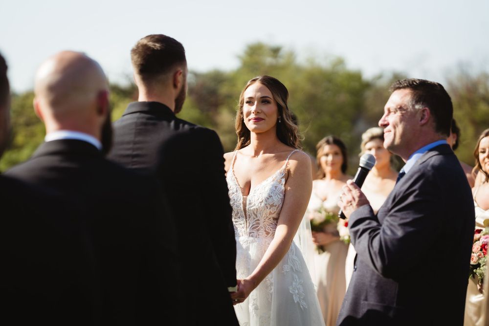 Wedding ceremony takes place outdoors on a bright sunny day with attendees gathered to witness the special moment.