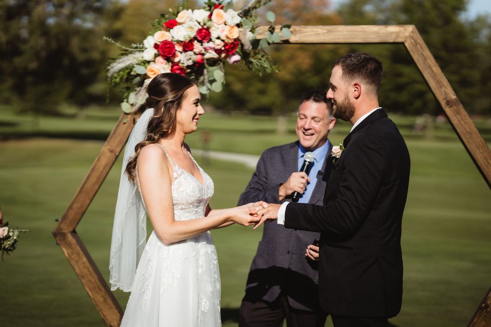 Outdoor wedding ceremony under a hexagonal wooden arch with red and white floral arrangements.