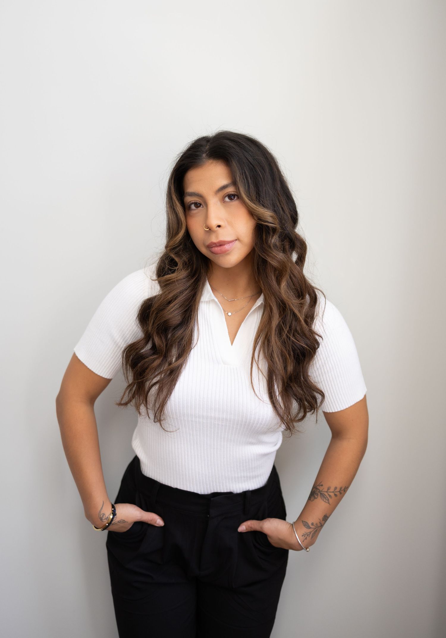 Young woman with long wavy brunette hair poses confidently in white ribbed top and black pants against neutral background.