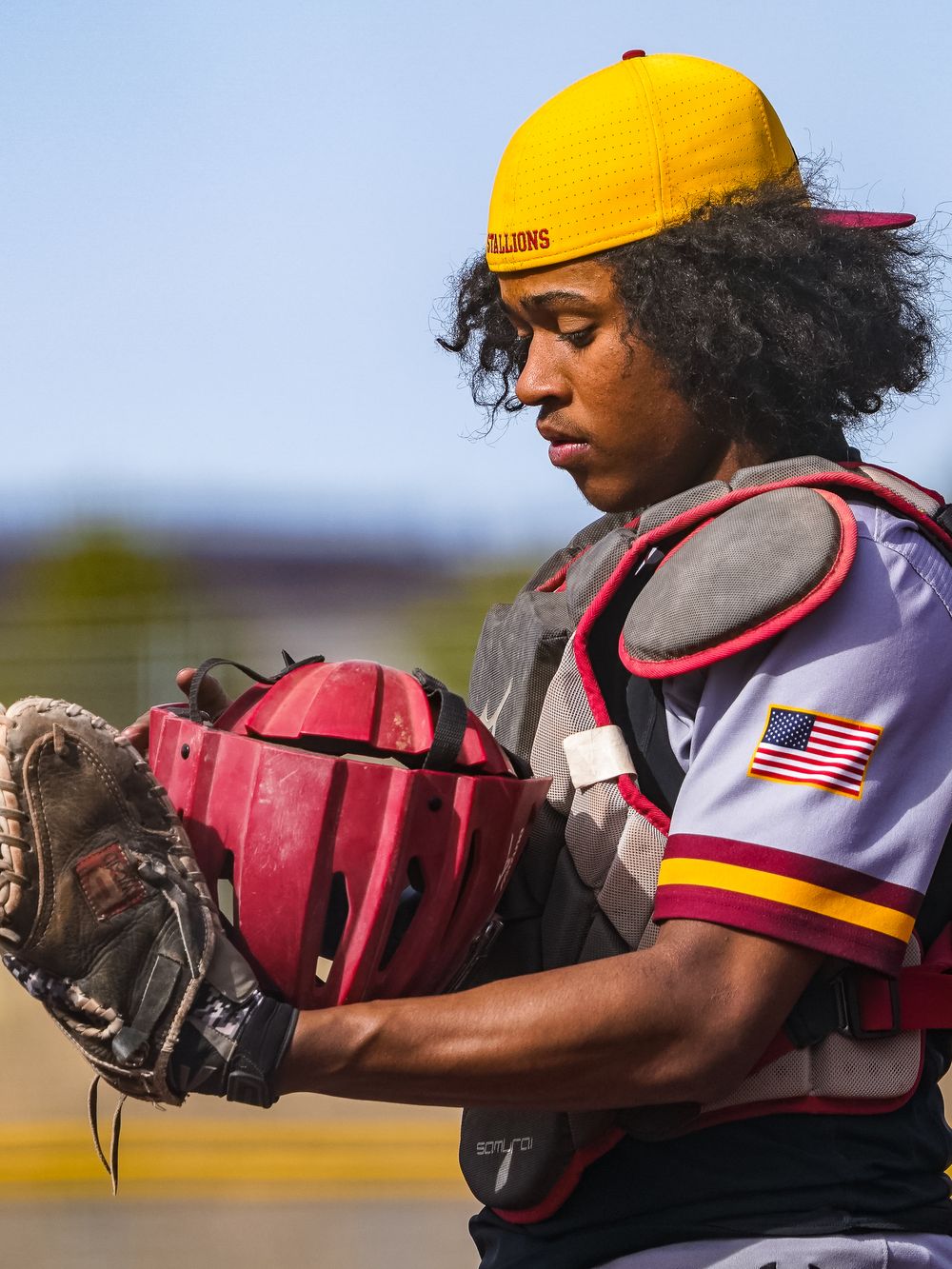 Young baseball catcher wearing yellow cap and protective gear examines mitt during game.