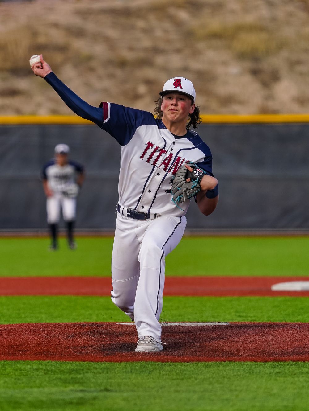Baseball pitcher in white uniform delivers pitch from mound during game on artificial turf field.