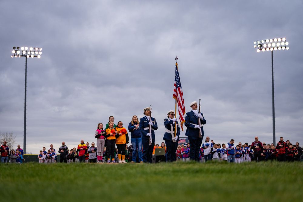 Color guard stands with American flag during outdoor sports event with stadium lights and crowd in background.