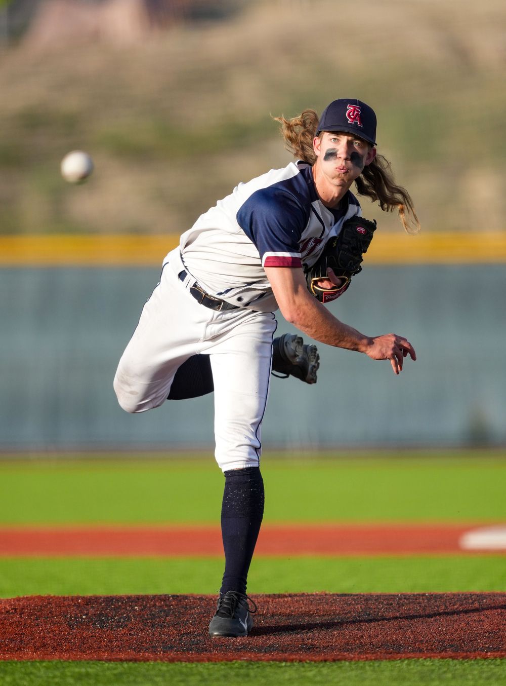 Baseball pitcher in navy uniform delivers pitch from mound during game on sunny day.