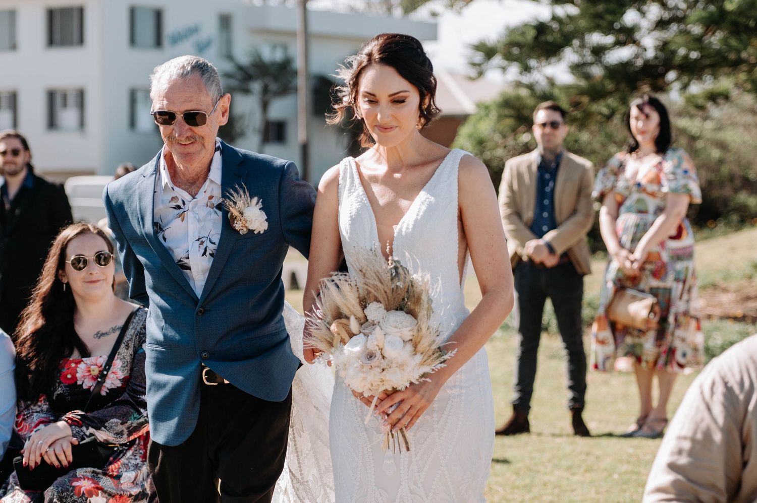 Outdoor coastal wedding ceremony with guests gathering on grass overlooking the ocean.