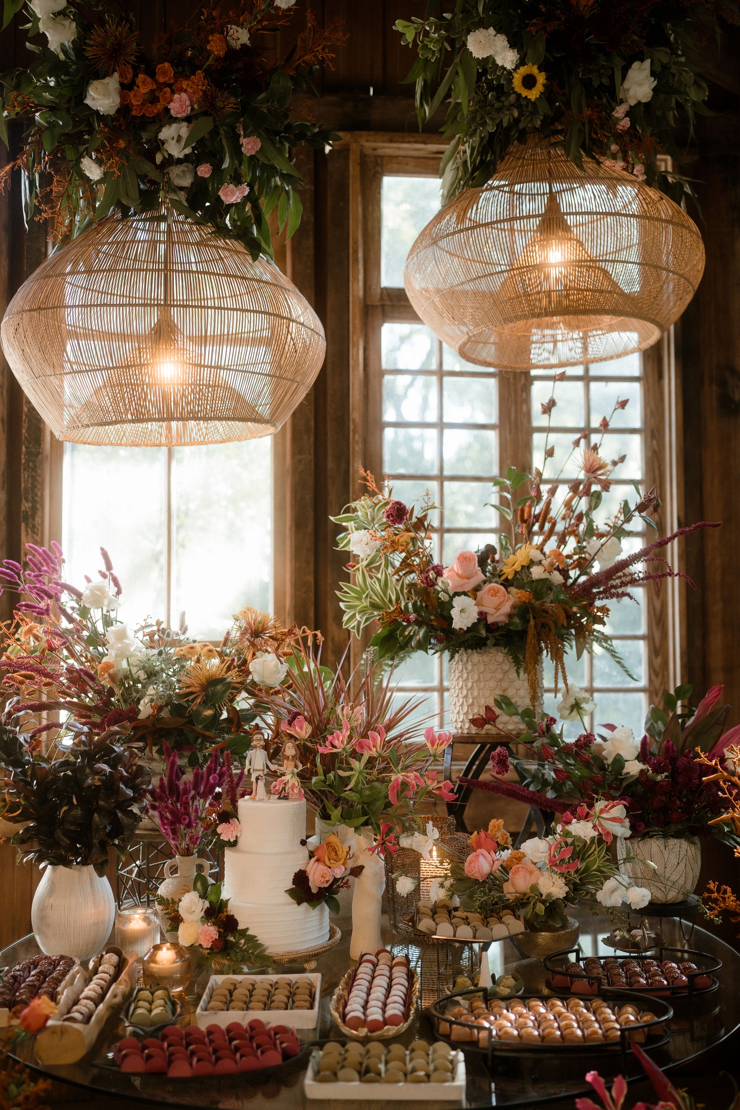 Elegant dessert table with floral arrangements and rattan pendant lights in a sunlit rustic venue.