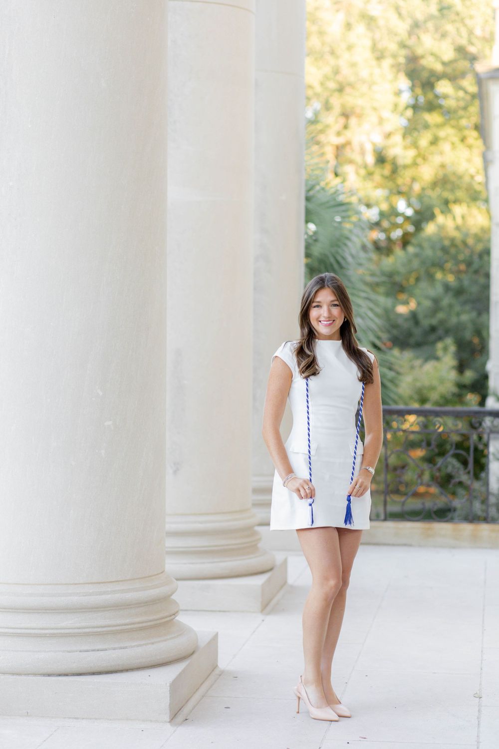 Graduate poses elegantly in white dress between classical columns on campus.