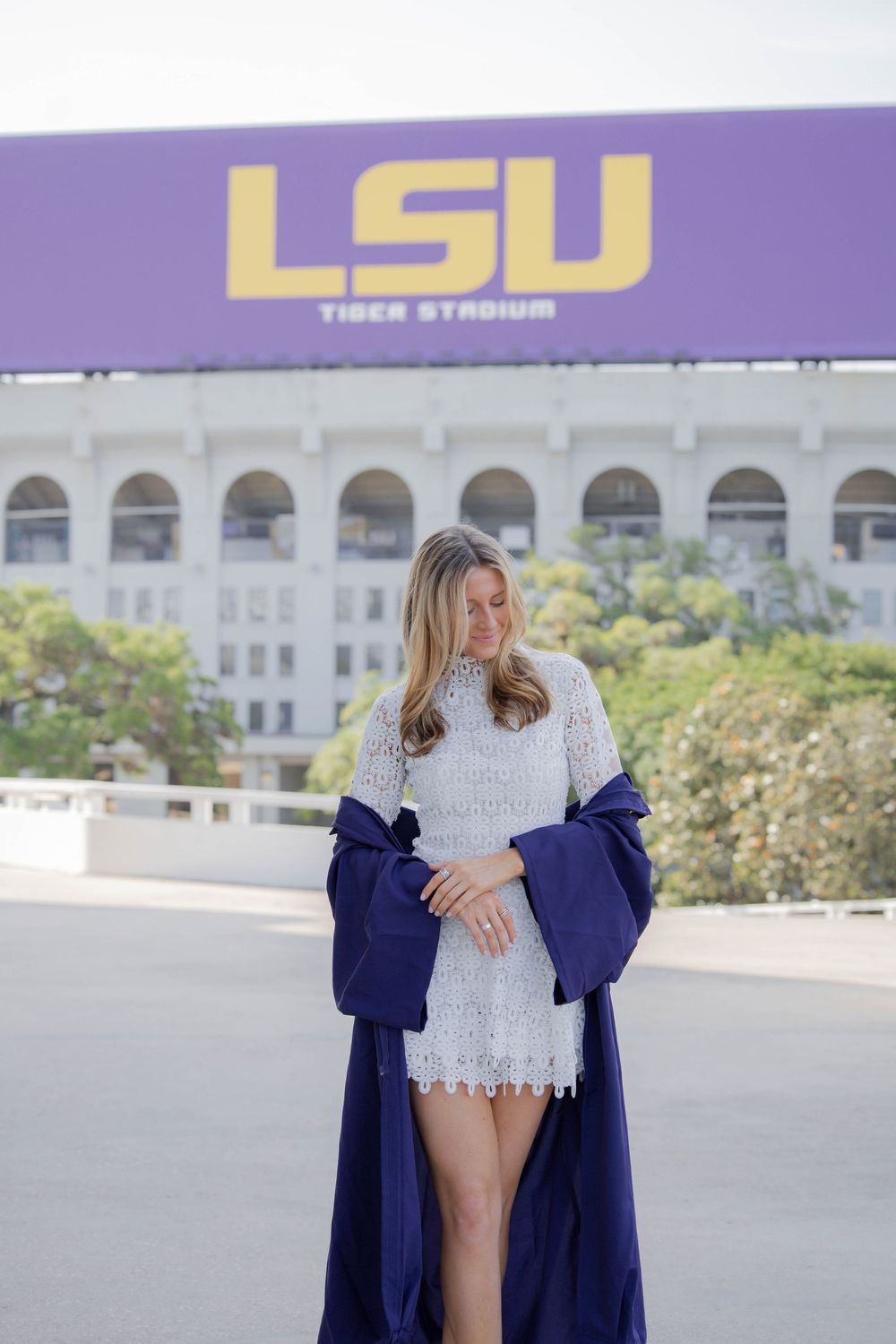 Person wearing blue graduation gown and white dress poses for photos outside of LSU Tiger Stadium.