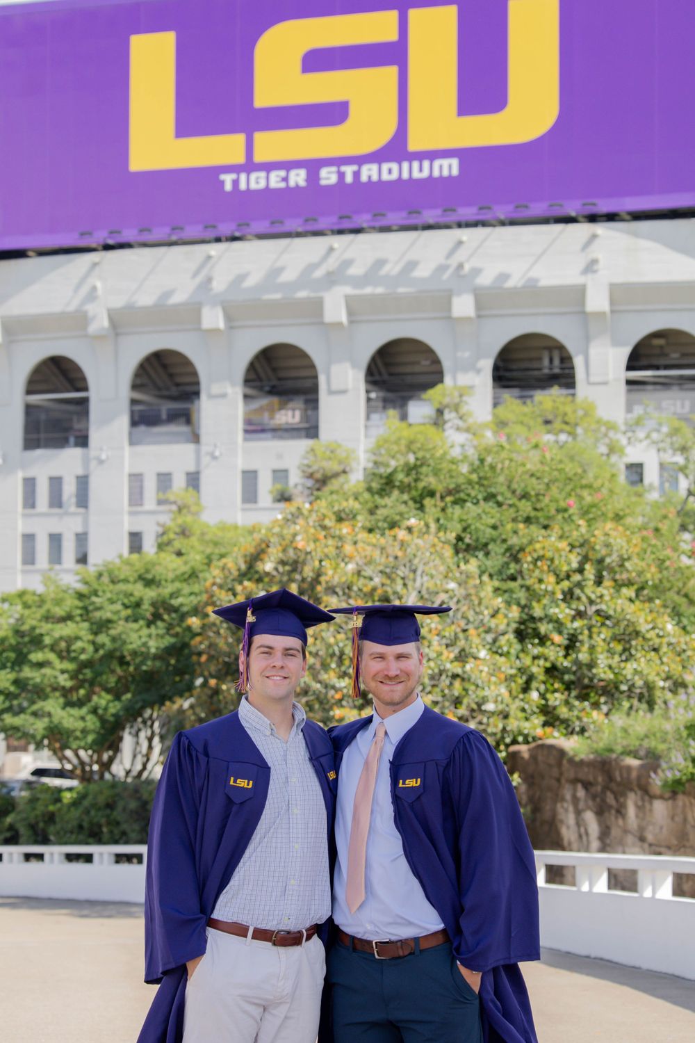Two graduates in LSU purple and gold regalia pose together outside Tiger Stadium.