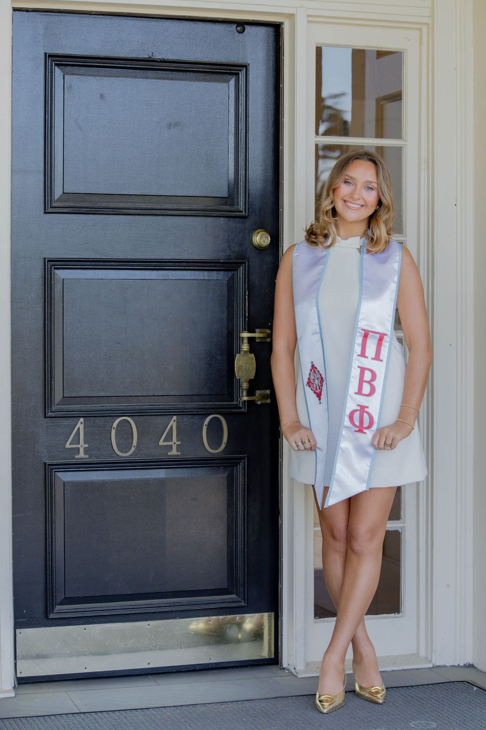 Someone in a light blue dress and graduation sash stands by a black door marked 4040, wearing gold shoes.