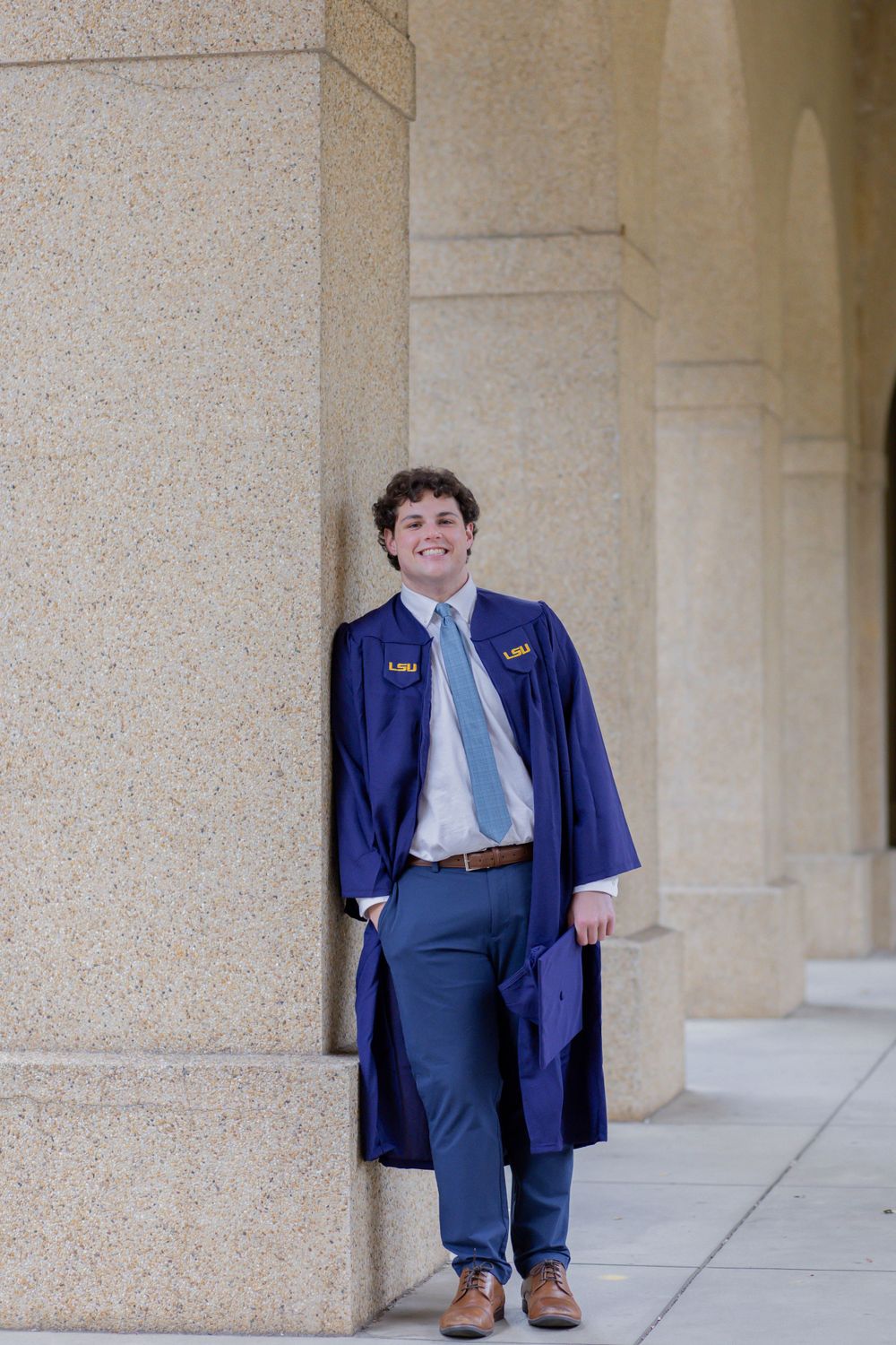A graduate in blue cap and gown stands in front of stone pillars on a university campus.