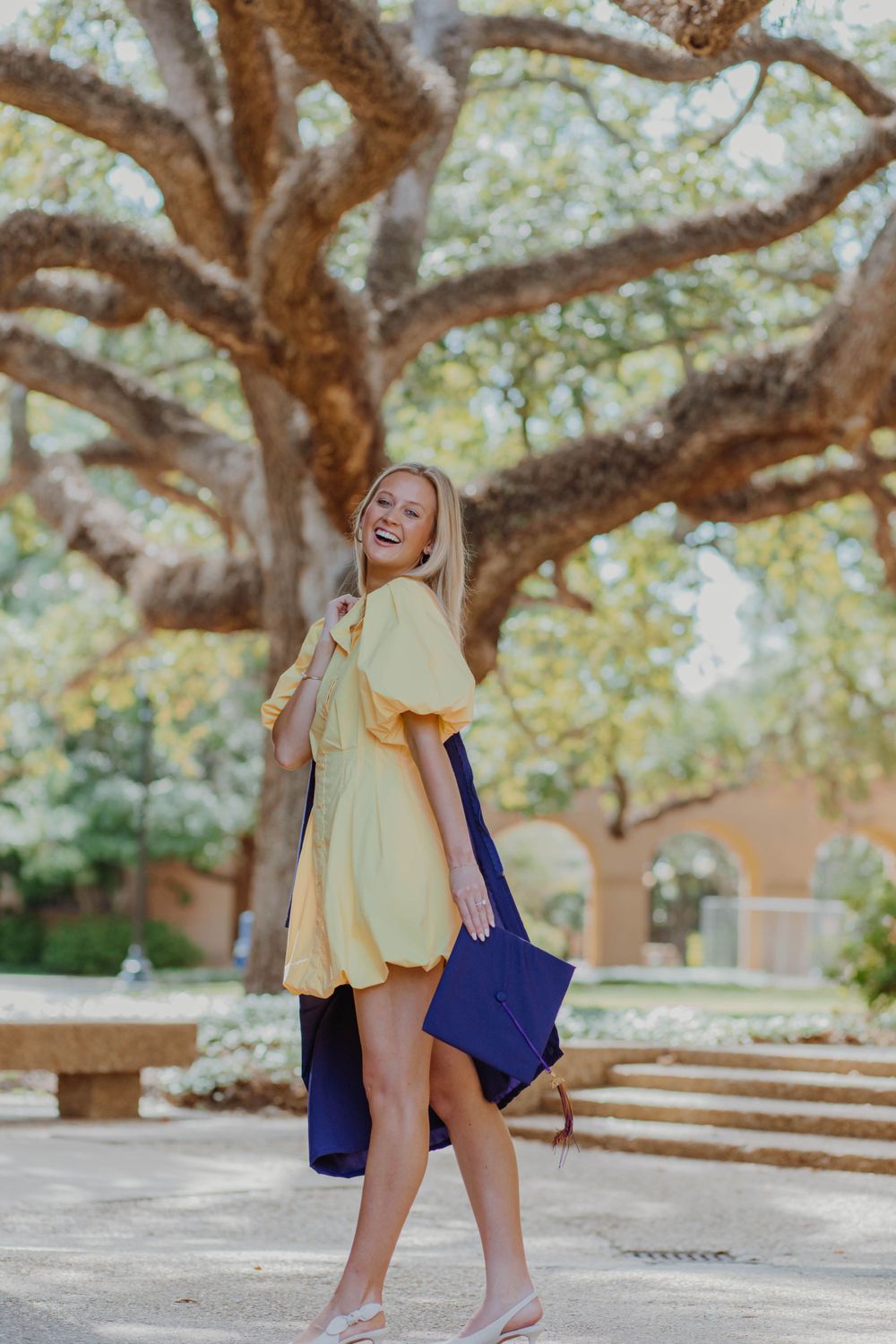 Graduate in yellow dress and purple graduation gown laughs joyfully under oak tree.